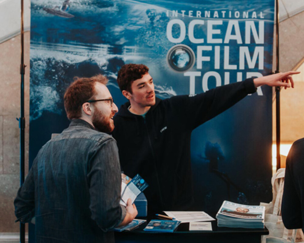 Stand der International Ocean Film Tour mit Mitarbeitern, die Besucher informieren und Magazine ausgeben.