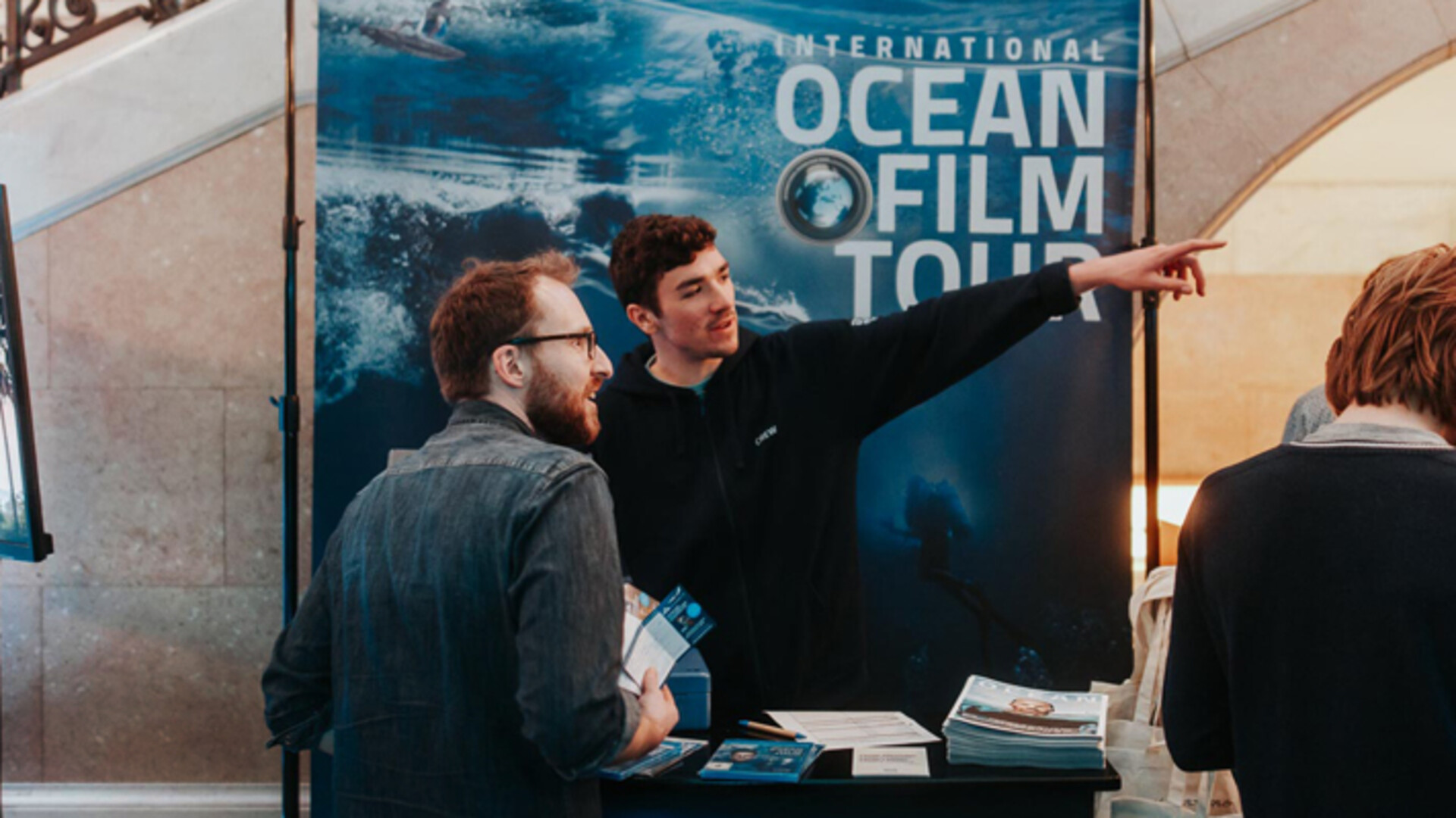 Stand der International Ocean Film Tour mit Mitarbeitern, die Besucher informieren und Magazine ausgeben.