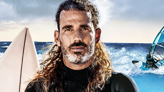 A man with long curly brown hair looks directly into the camera and is wearing a wetsuit with his arms crossed in front of his chest.