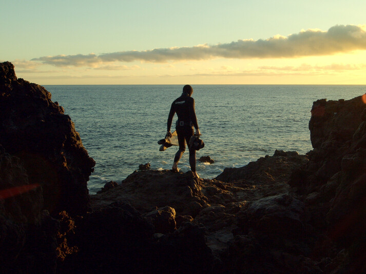 Plongeur en combinaison debout sur des rochers au bord de la mer au coucher du soleil, tenant son équipement de plongée.