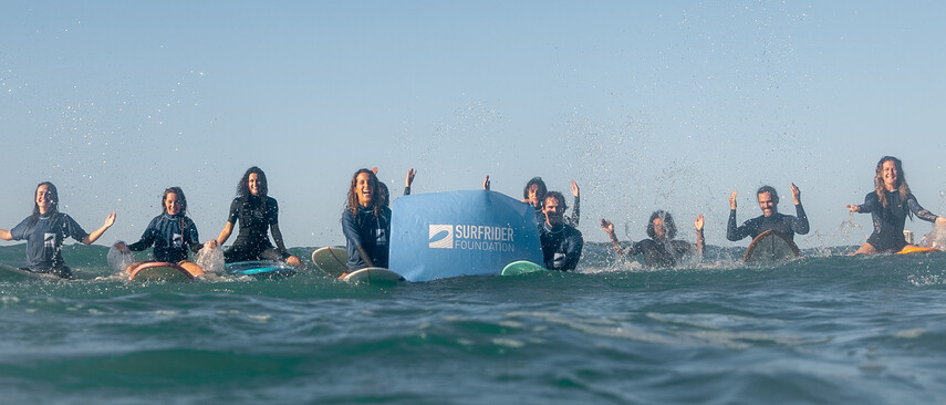 Eine Gruppe sitzen auf Surfbrettern und halten ein blaues Banner mit dem Surfrider Foundation Logo hoch